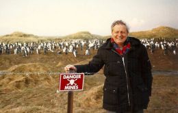 Harold Briley en una postal de las Falklands, junto a un cartel advirtiendo de un campo minado y en el fondo una colonia de pingüinos